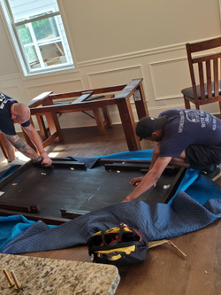 Two men assembling a dark wooden table in a room with hardwood floors.