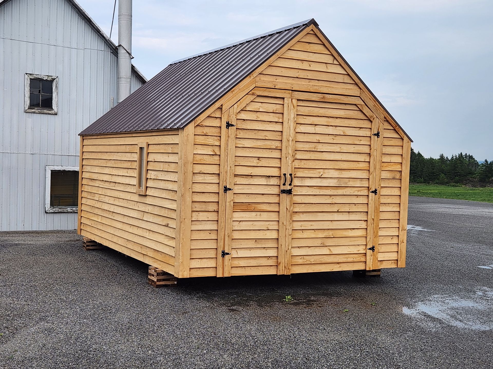 Un petit hangar en bois au toit marron est garé devant un bâtiment blanc.