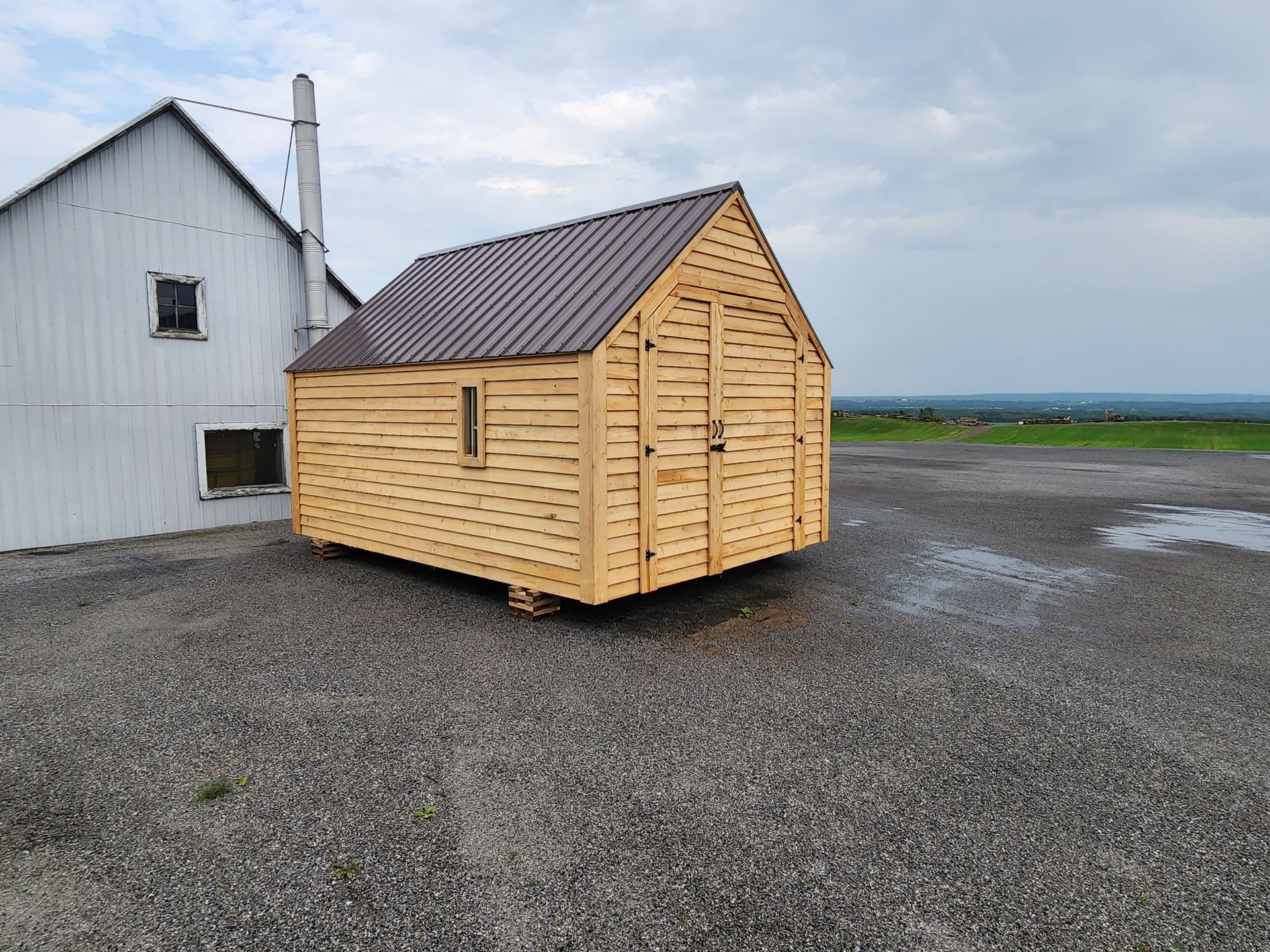 Un petit hangar en bois se trouve sur un terrain en gravier devant une grange blanche.