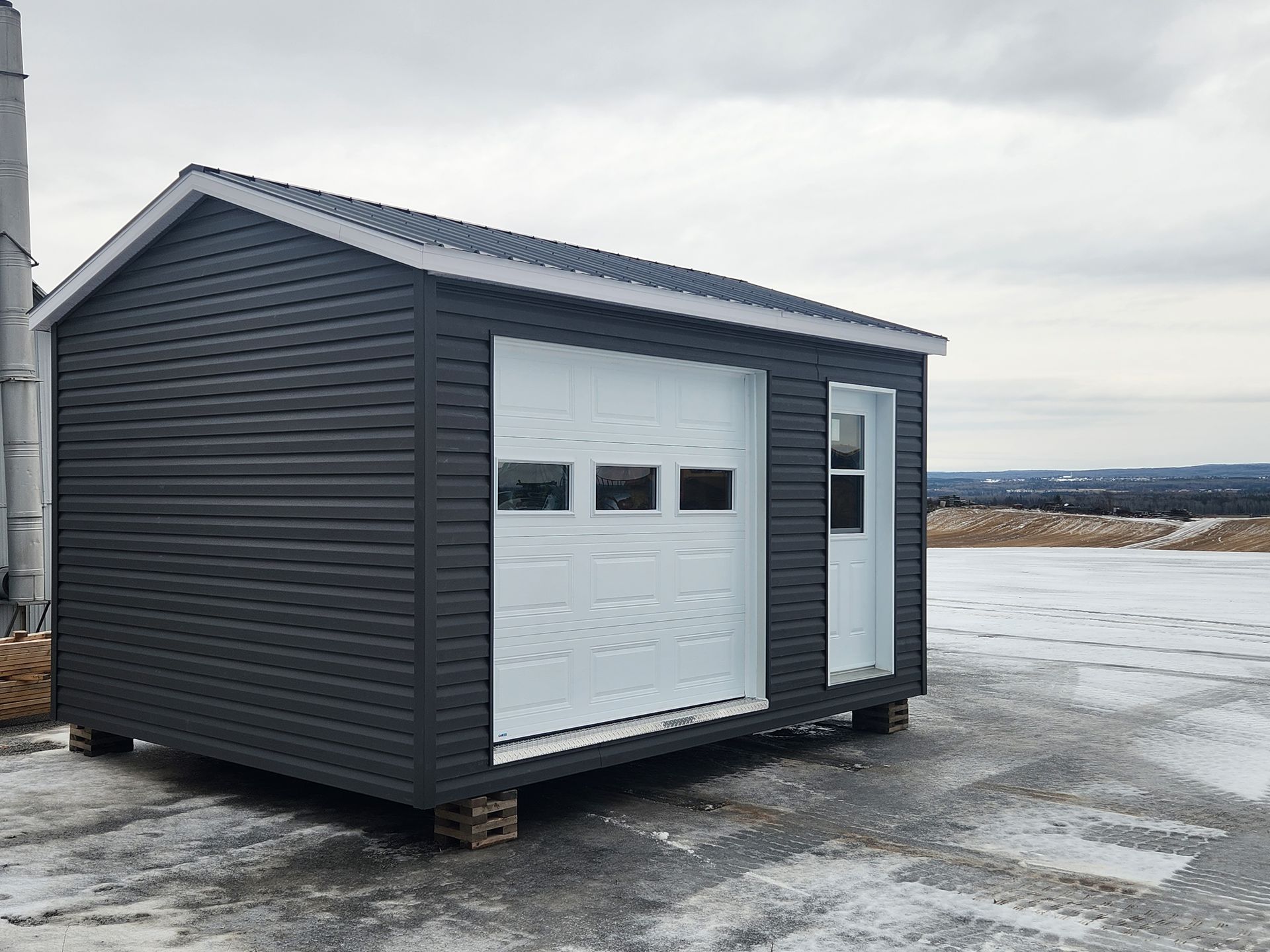 Un hangar noir avec une porte de garage blanche est assis dans la neige.