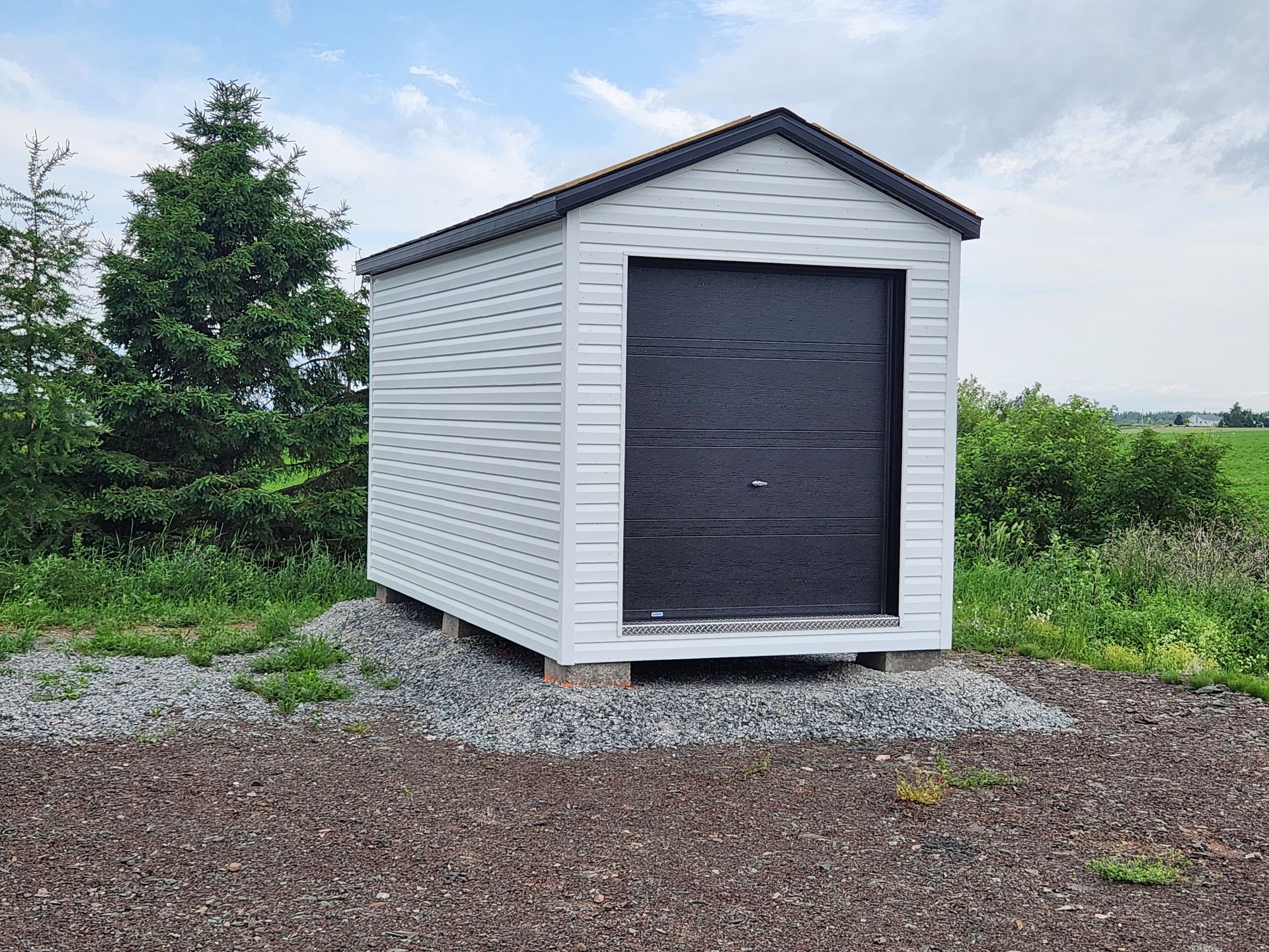 Un petit hangar blanc avec une porte de garage noire se trouve au milieu d'un champ.
