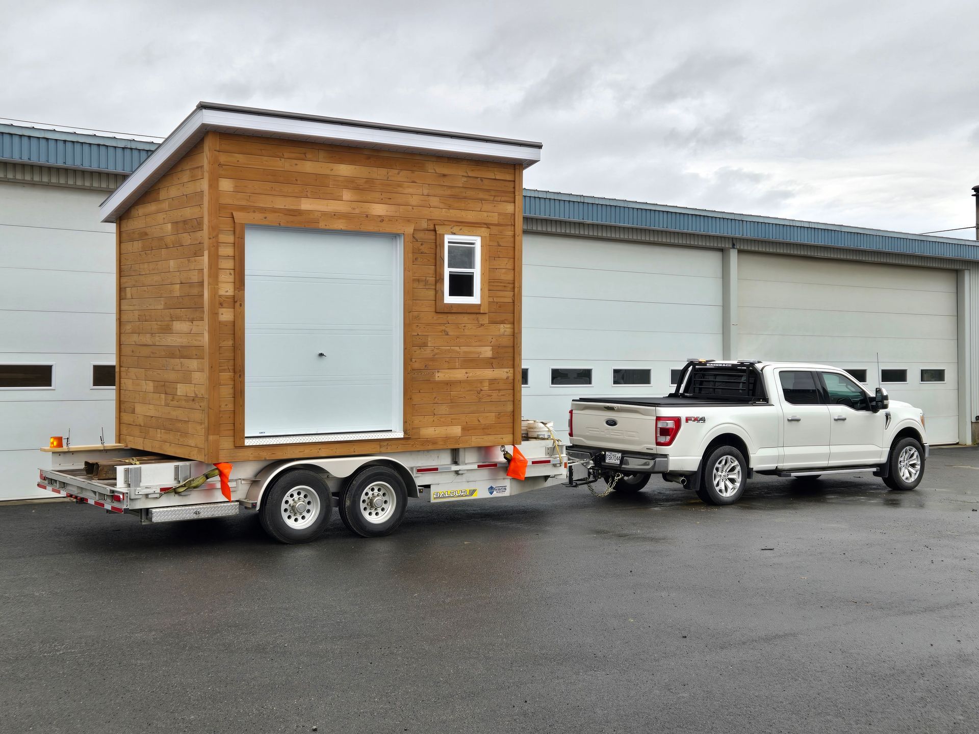 Un camion blanc tracte un petit hangar en bois sur une remorque.