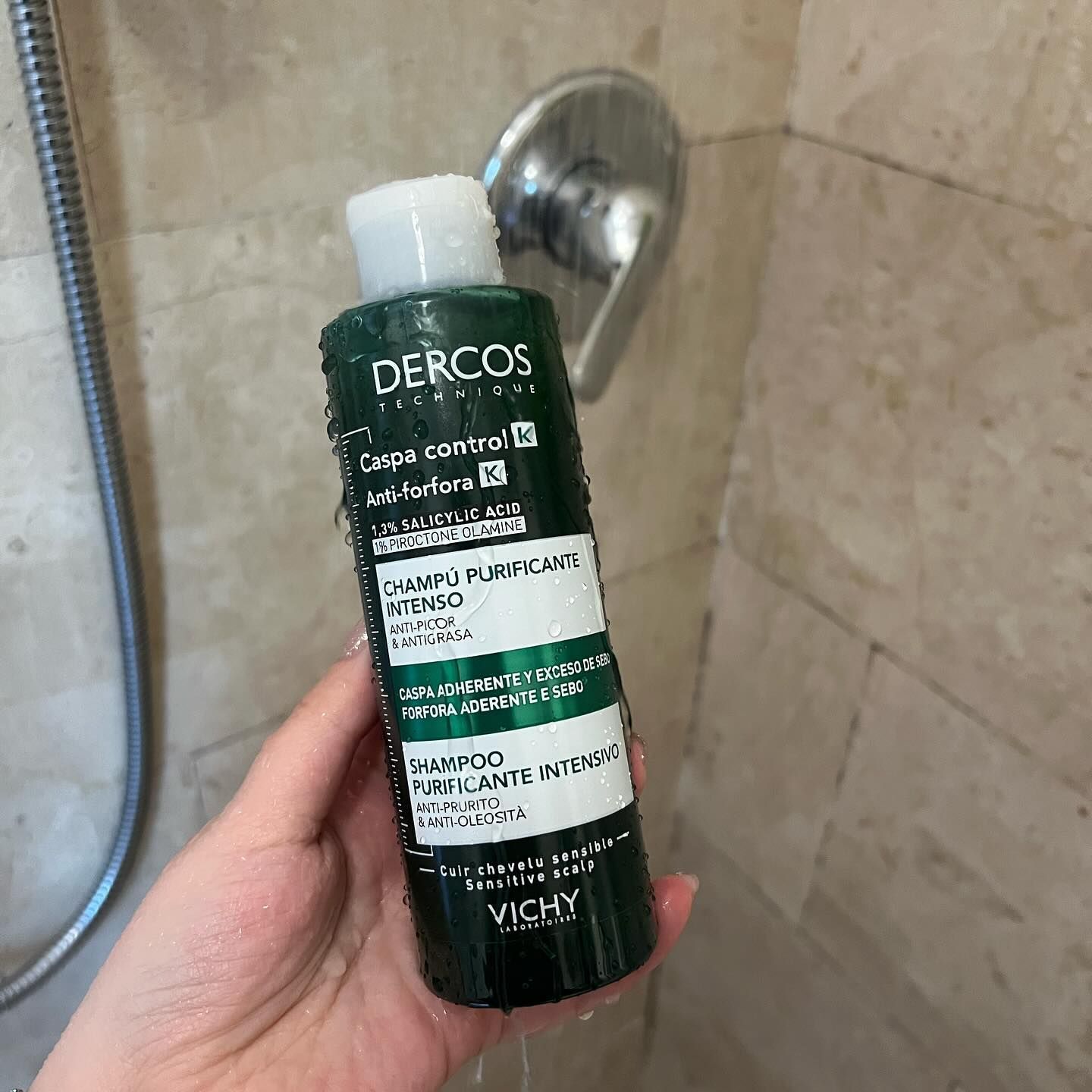 Mano che tiene una bottiglia di shampoo Derkos verde e bianca sotto la doccia con acqua corrente.