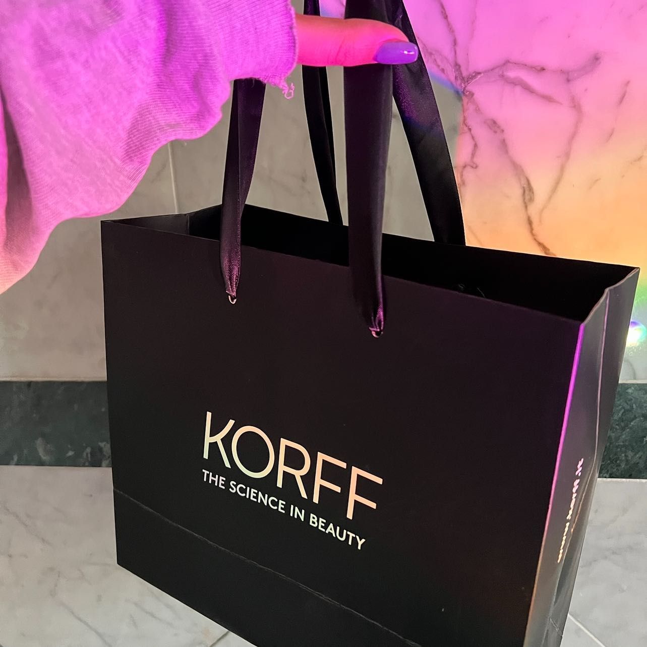 Borsa della spesa nera KORFF con scritta dorata, tenuta da una persona con unghie viola, sfondo rosa.
