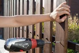A hand uses a cordless drill to fasten a wooden board to a vertical slat fence outdoors.