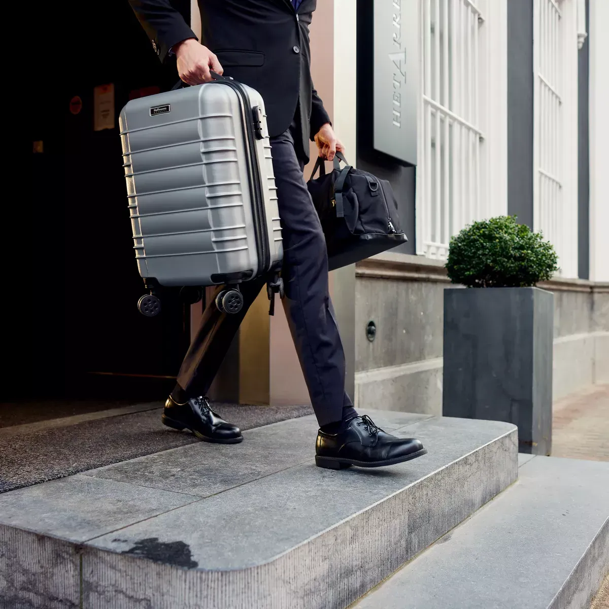 Man in black suit stepping out of a building, carrying a silver suitcase and a black bag.