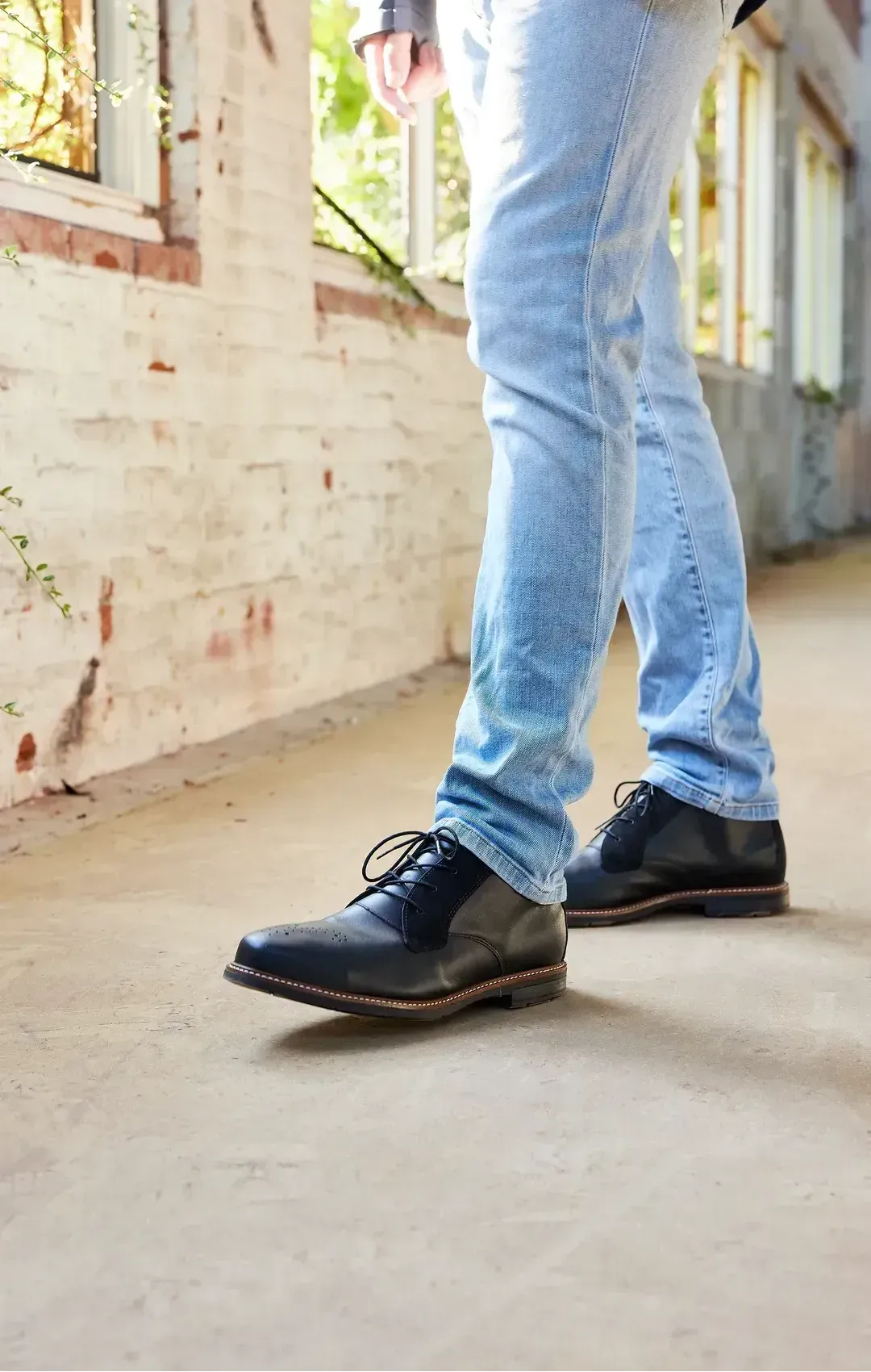 Person walking in a building, wearing blue jeans and black leather shoes. The setting has a concrete floor and brick walls.