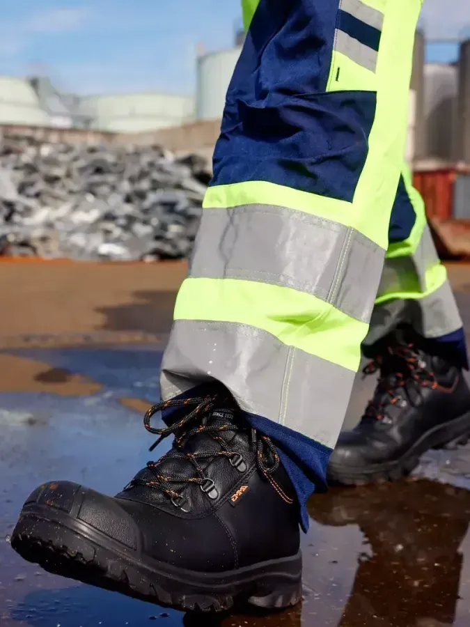 A person wearing safety pants and boots is walking in a puddle.