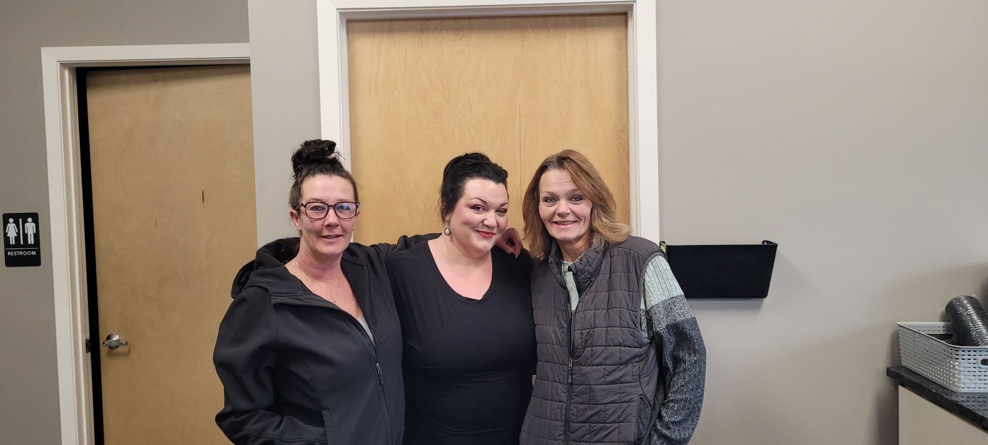 Three people standing together and smiling in an office setting with light walls and two doorways.