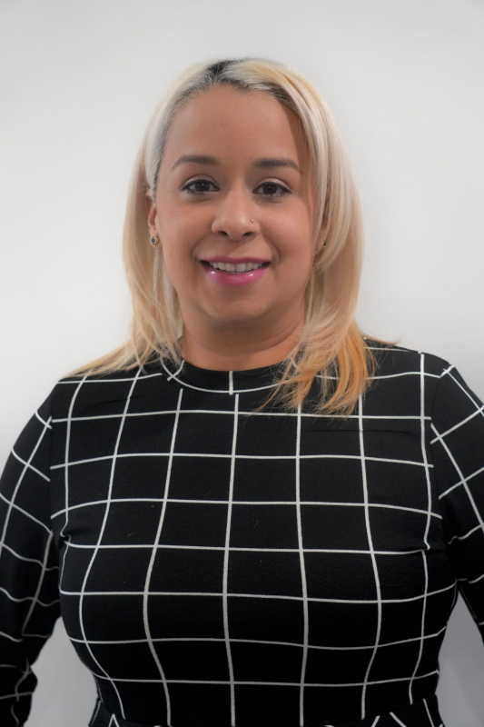 A smiling individual with light blonde hair wearing a black shirt with a white grid pattern against a white background. Carla, Office Manager
