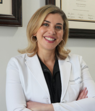 A smiling professional in a white lab coat with crossed arms, standing in an office with framed certificates on the wall. Dr. Habib Mariam