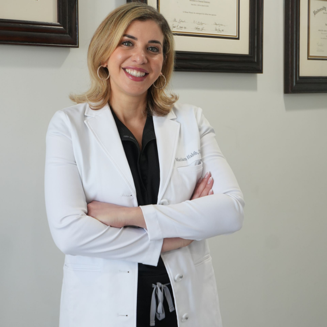 A smiling professional in a white lab coat with crossed arms, standing in an office with framed certificates on the wall. Dr. Habib Mariam