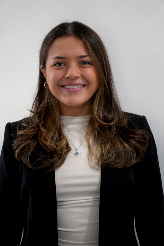 A smiling person with long brown hair wearing a white top and a black blazer against a plain white background. Dr. Paredes