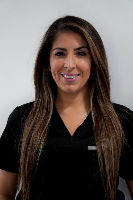 A person with long brown hair, wearing a black V-neck top, smiling against a plain white background. Sofia, Dental Hygienist