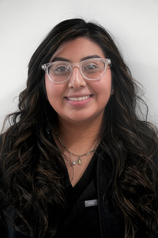 A smiling person with long dark wavy hair wearing clear-framed glasses and a dark scrub top against a white background. Stephanie, Dental Assistant