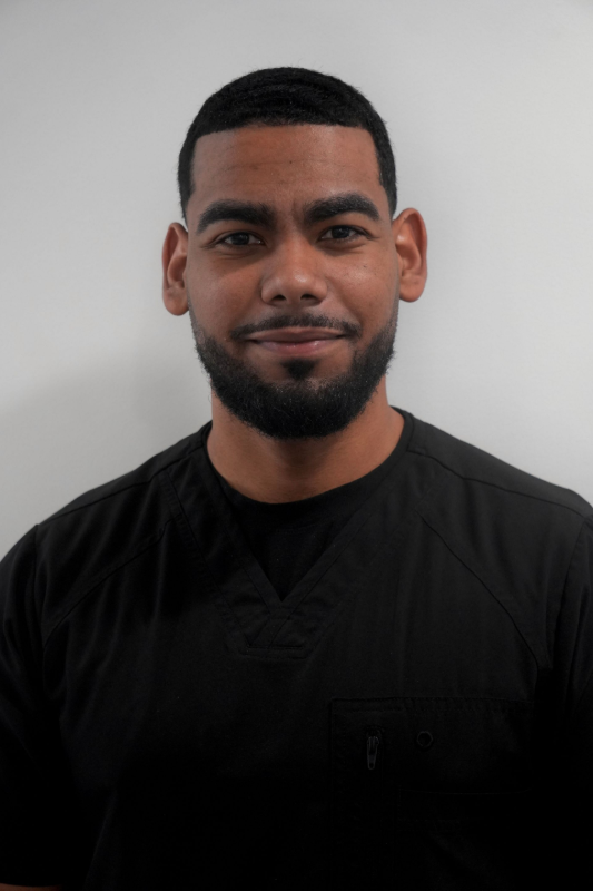 A man wearing a black scrub top looks toward the camera against a plain, light-colored background. Steven, Dental Assitant.