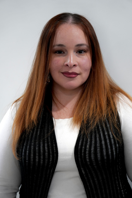 A person with long, reddish-brown hair wearing a white top and a black textured vest, posing against a plain backdrop. Wandalee, Front Desk Receptionist