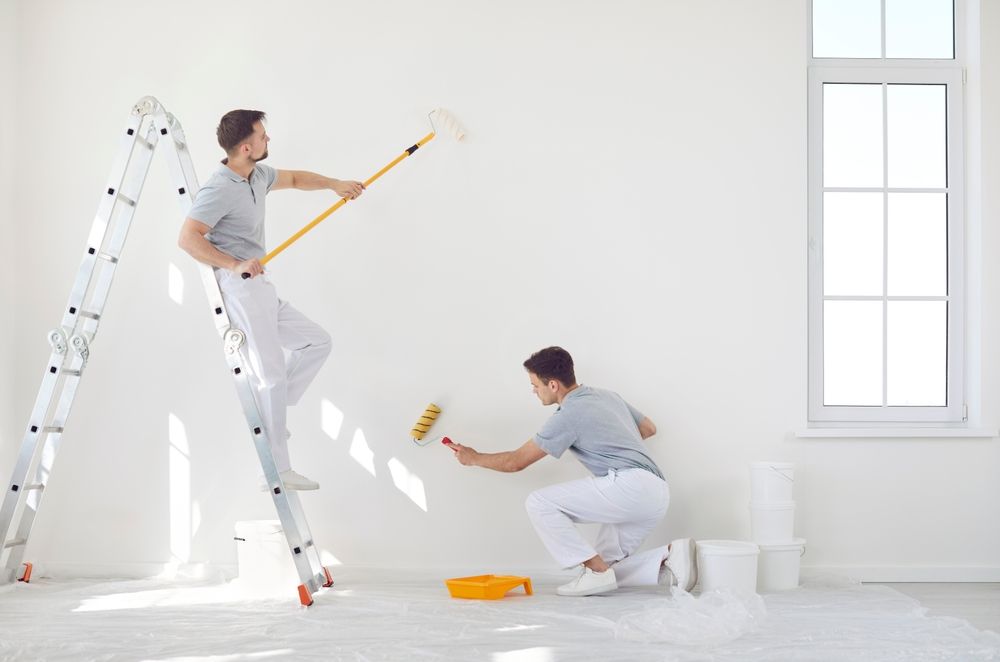 Two painters in gray shirts and white pants painting a white wall with rollers and a ladder.