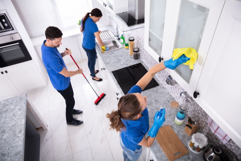 Three people wearing blue shirts cleaning a kitchen; one sweeping, one washing dishes, and one wiping cabinets.