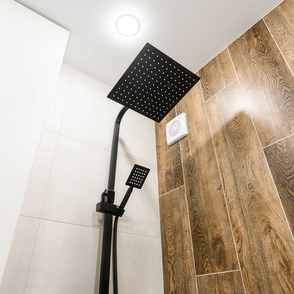 Black showerhead with handheld shower in a shower stall with wood-look tile and white tile walls.