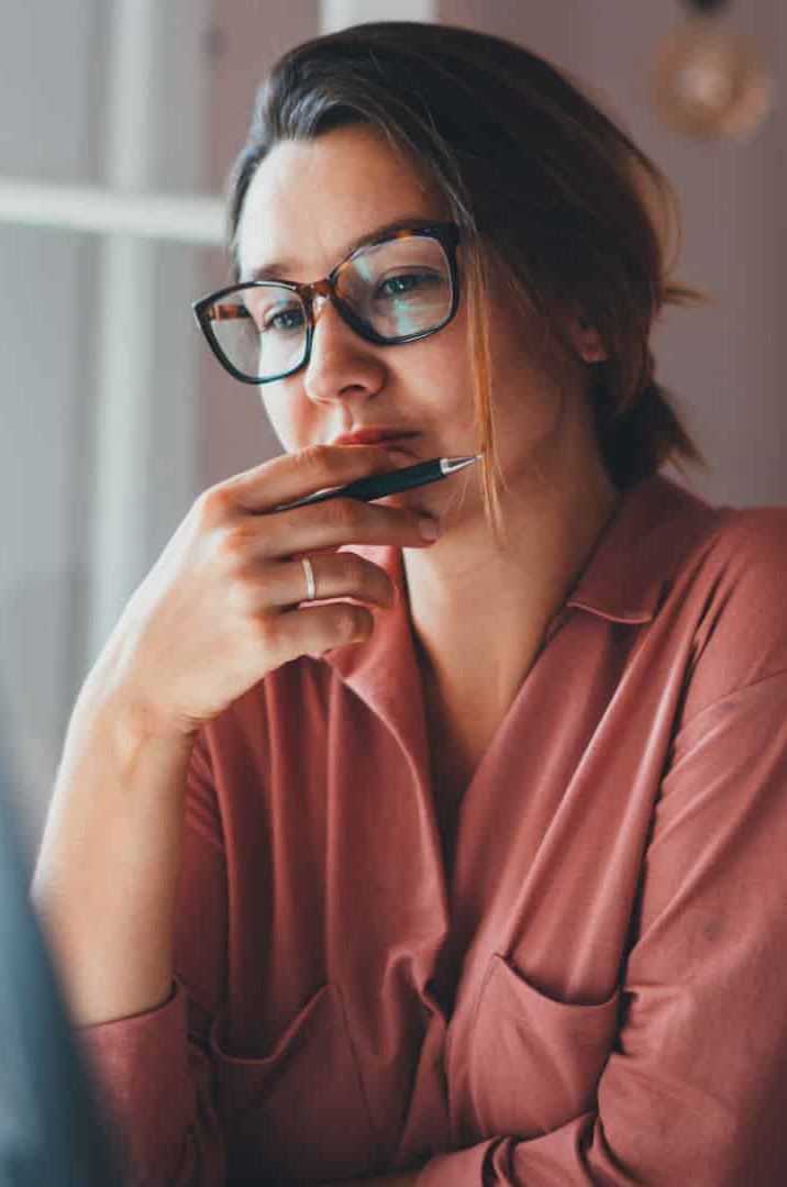 A woman wearing glasses is holding a pen to her mouth.
