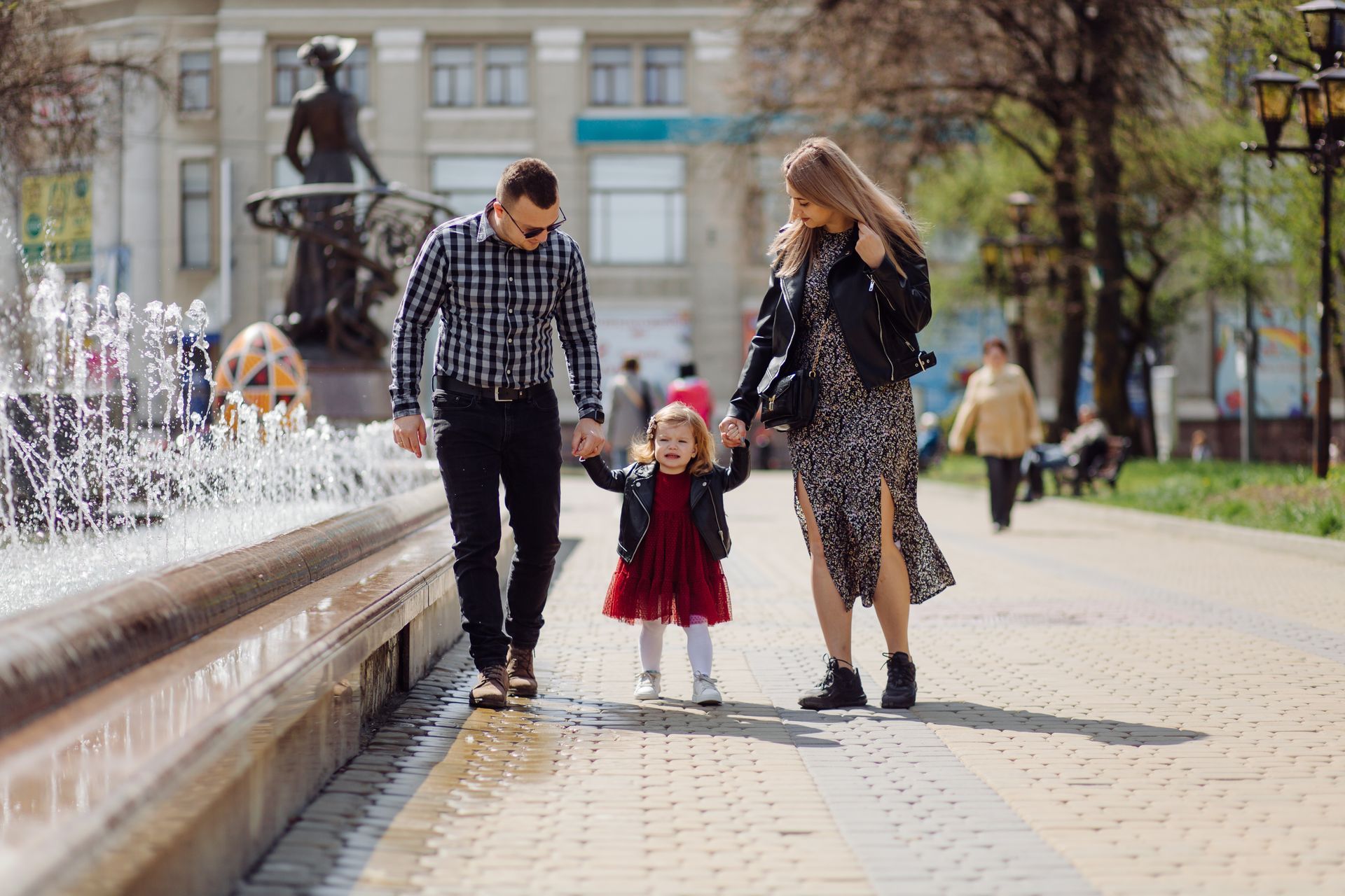 Family of three walking near a fountain; the child holds parents' hands, smiling.