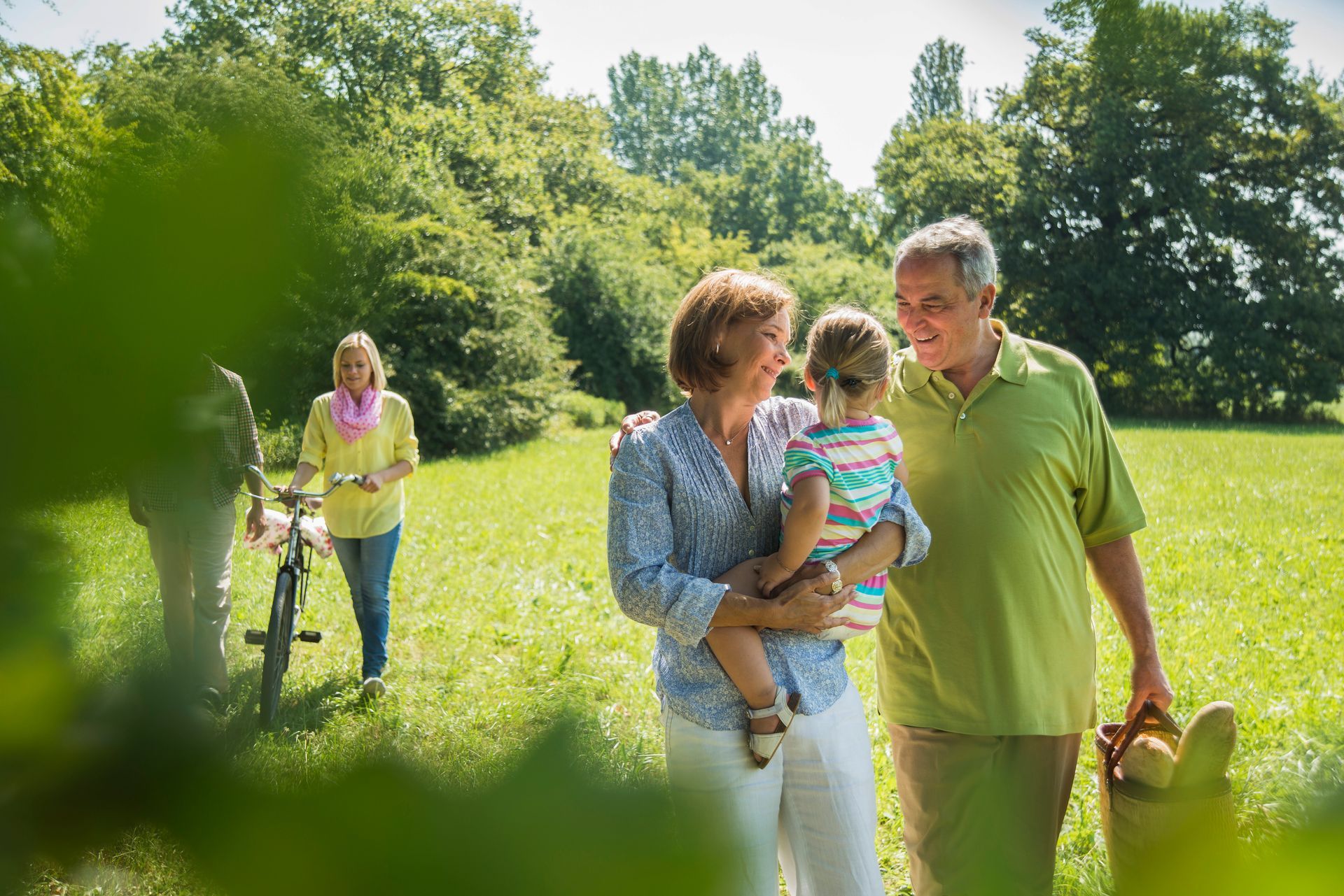 Family picnicking in a sunny park; grandparents hold a child, other woman walks with a bicycle.