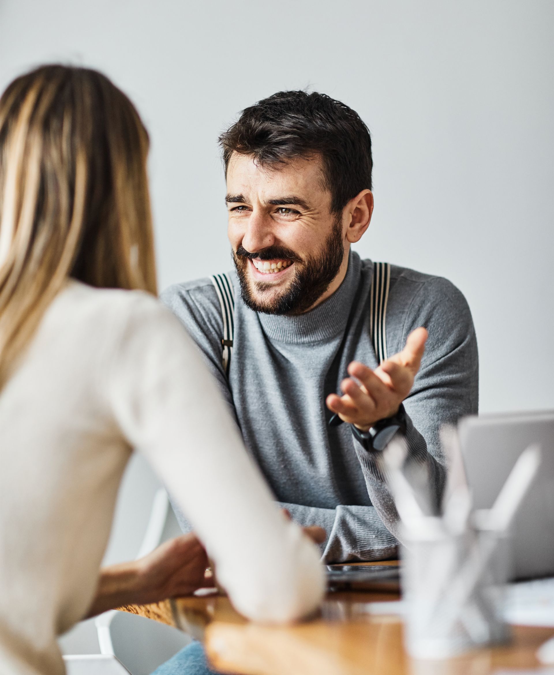 Man with beard smiles and gestures while talking to a person at a table, near a laptop and pens.
