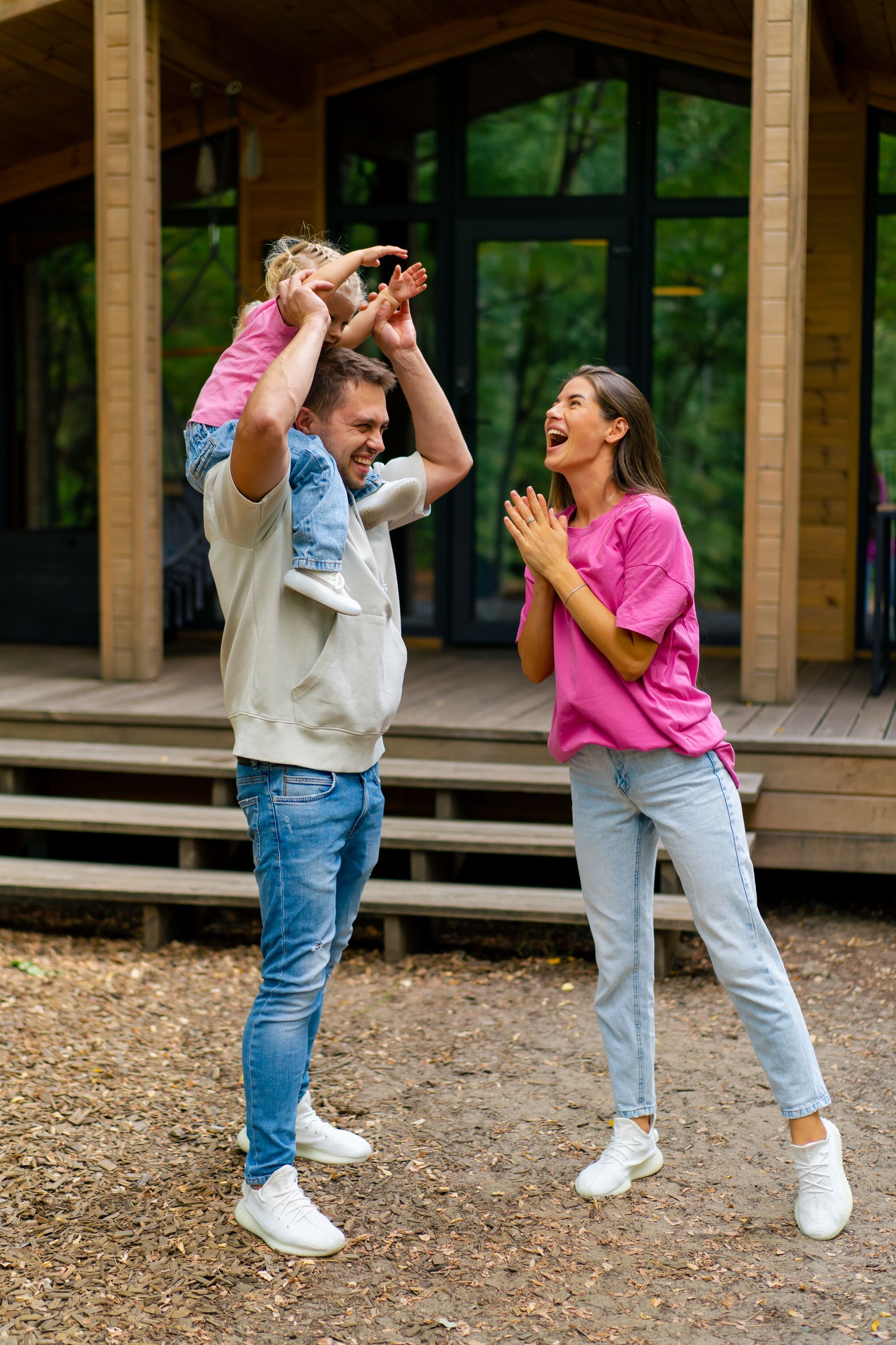 Man carrying child on shoulders, woman watching with excitement in front of a wooden house.