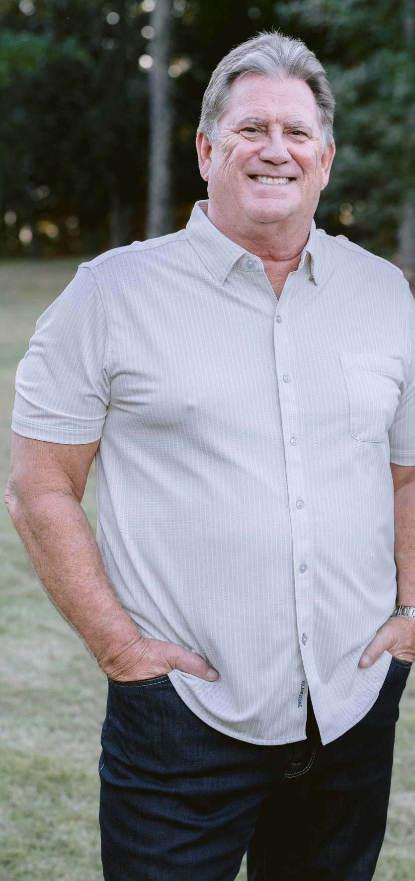 Man in a button-down shirt and jeans, smiling, standing outside with hands in pockets.