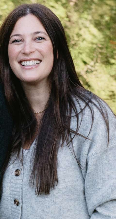 Woman with long brown hair smiles, wearing a gray cardigan, against a blurred green backdrop.