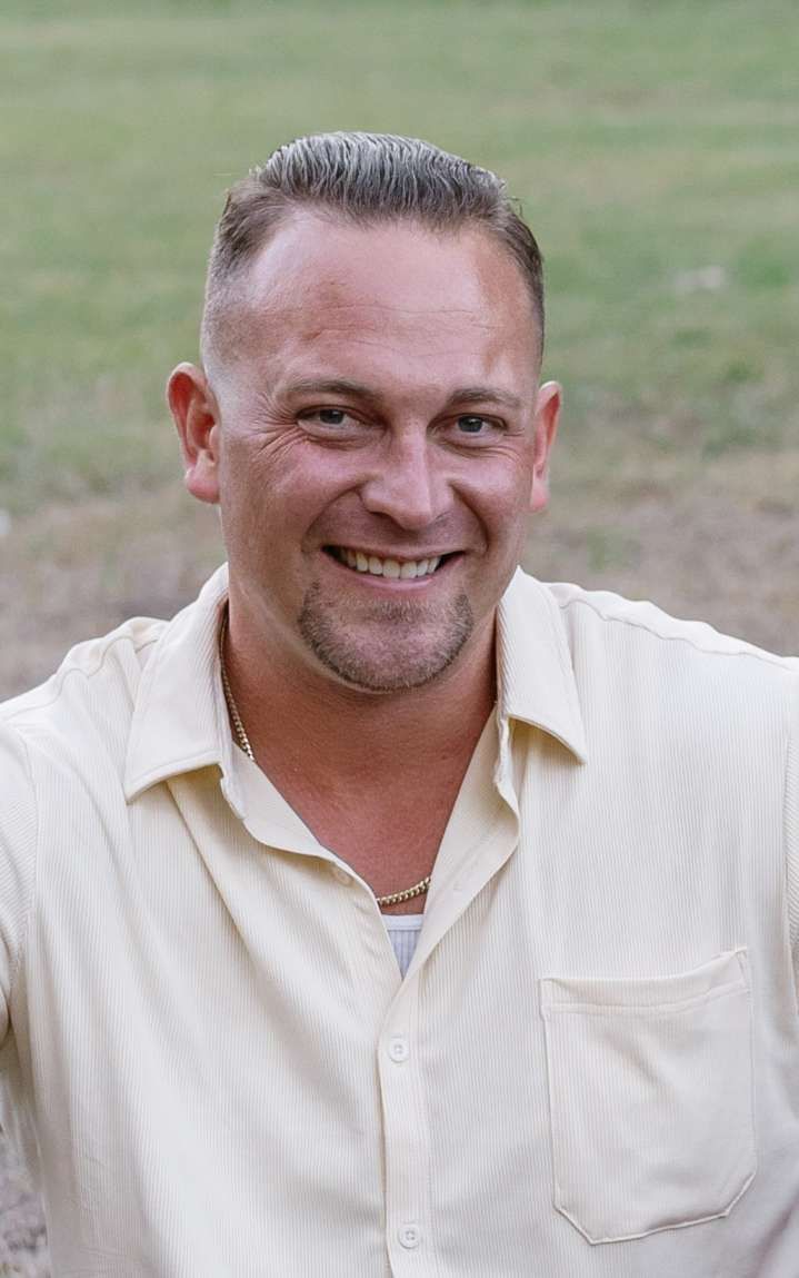Man with short hair and a goatee smiles wearing a light yellow button-up shirt outdoors.