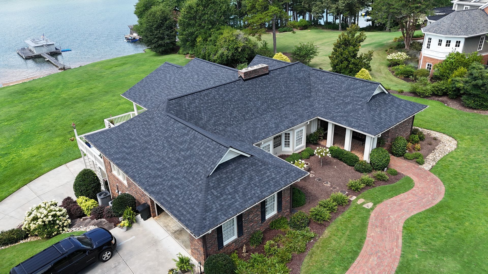 House with dark roof near a lake, surrounded by green lawn and a brick walkway.