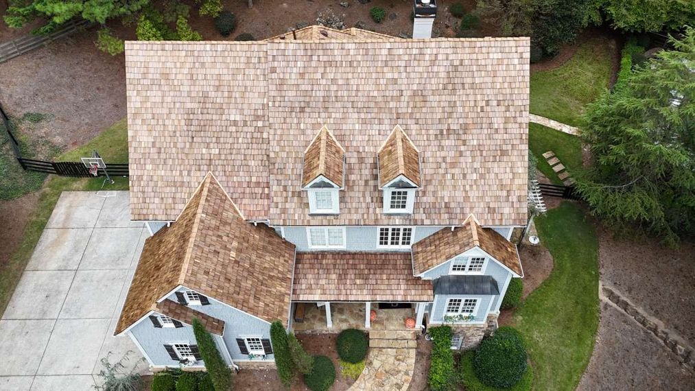 Aerial view of a gray house with a wood shingle roof, stone walkway, and lush surrounding trees.