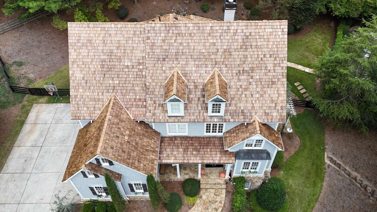 Aerial view of a gray house with a wood shingle roof, stone walkway, and lush surrounding trees.