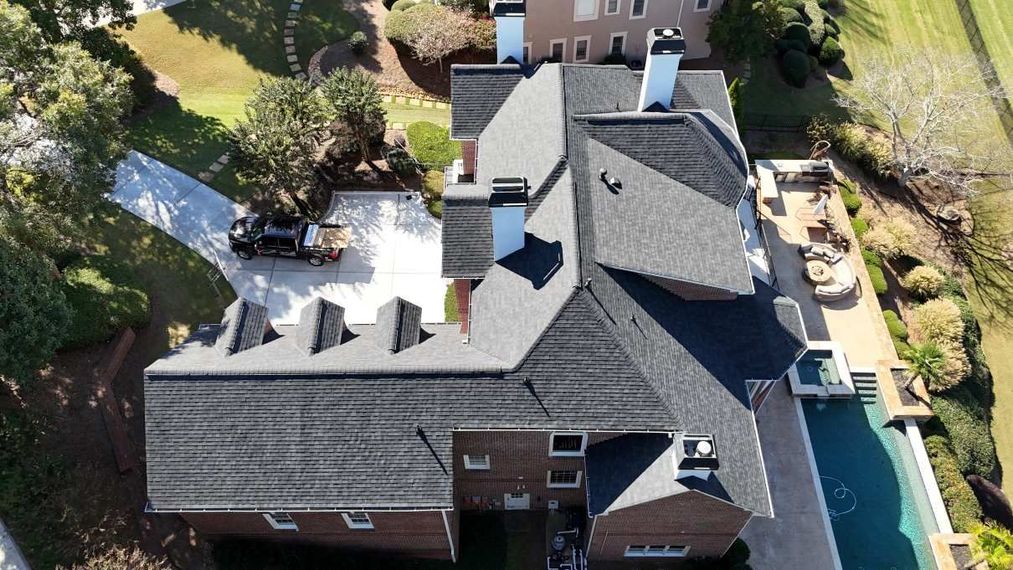 An aerial view of a brick house with a dark shingled roof, showing a driveway, chimney stacks, and a backyard pool area.