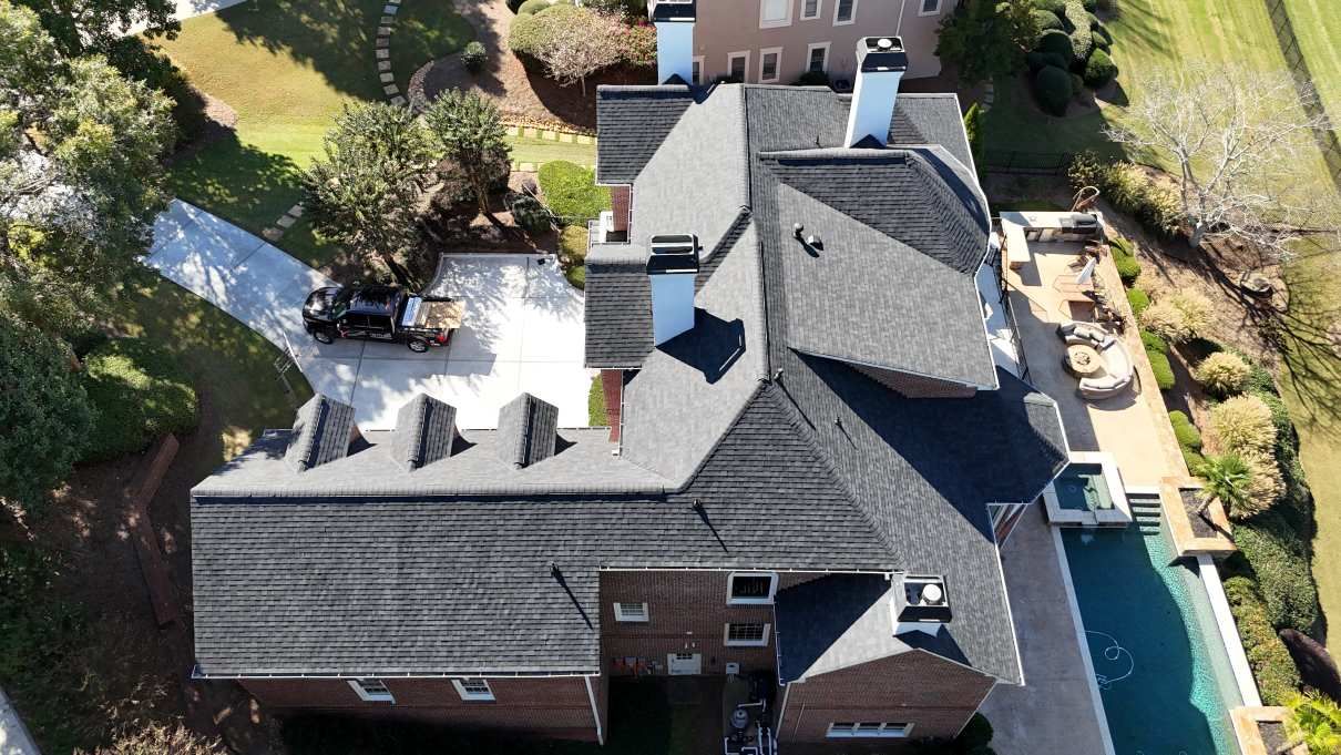 An aerial view of a brick house with a dark shingled roof, showing a driveway, chimney stacks, and a backyard pool area.