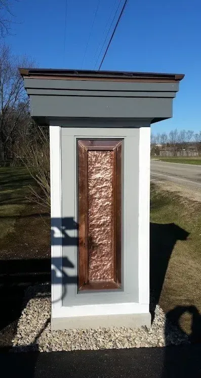 A copper-paneled pillar with a gray top and base, set in gravel next to a road, under a bright sky.