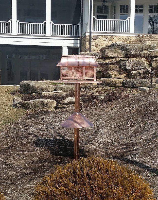 Copper birdhouse atop a pole, with a copper roof and a sunshade, in front of a stone wall and a house.