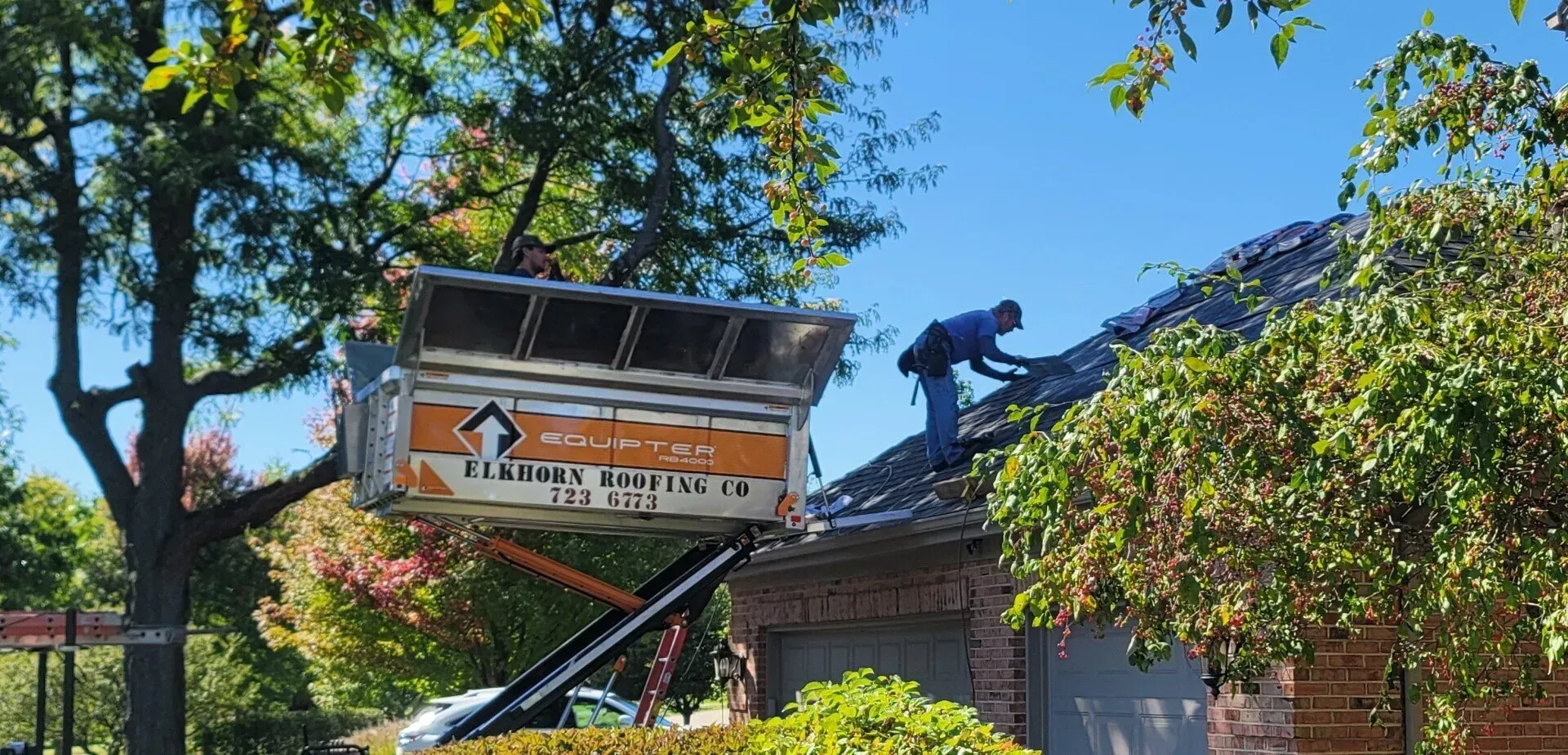 A roofer on a house roof is working near a dumpster being hoisted by a lift, sunny day. A roofer on a house roof is working near a dumpster being hoisted by a lift, sunny day.