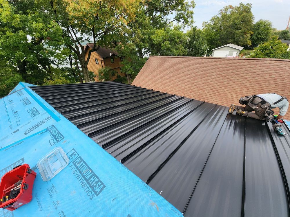 Roofer installing black metal panels on a roof covered in blue underlayment, with a brown roof in the background.