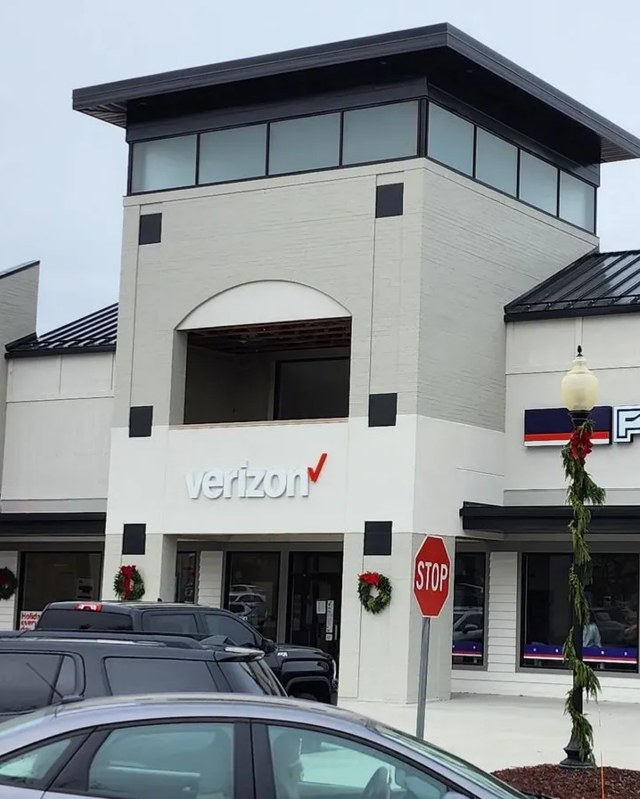 Verizon store exterior with gray brick facade, open second story, and black roof. A stop sign and garland visible. Verizon store exterior with gray brick facade, open second story, and black roof. A stop sign and garland visible.