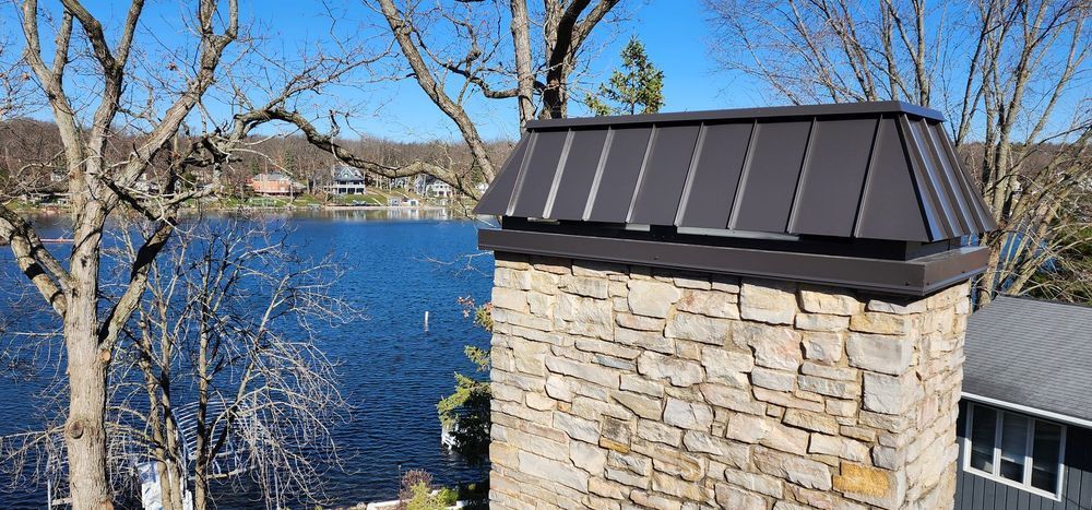 Stone chimney with dark metal cap against a lake and blue sky.