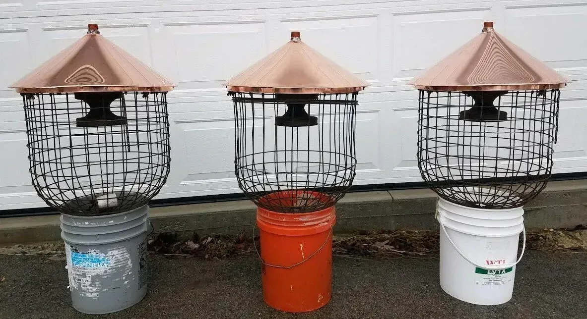 Three copper-topped bird feeders with wire cages sit on buckets, next to a white surface.