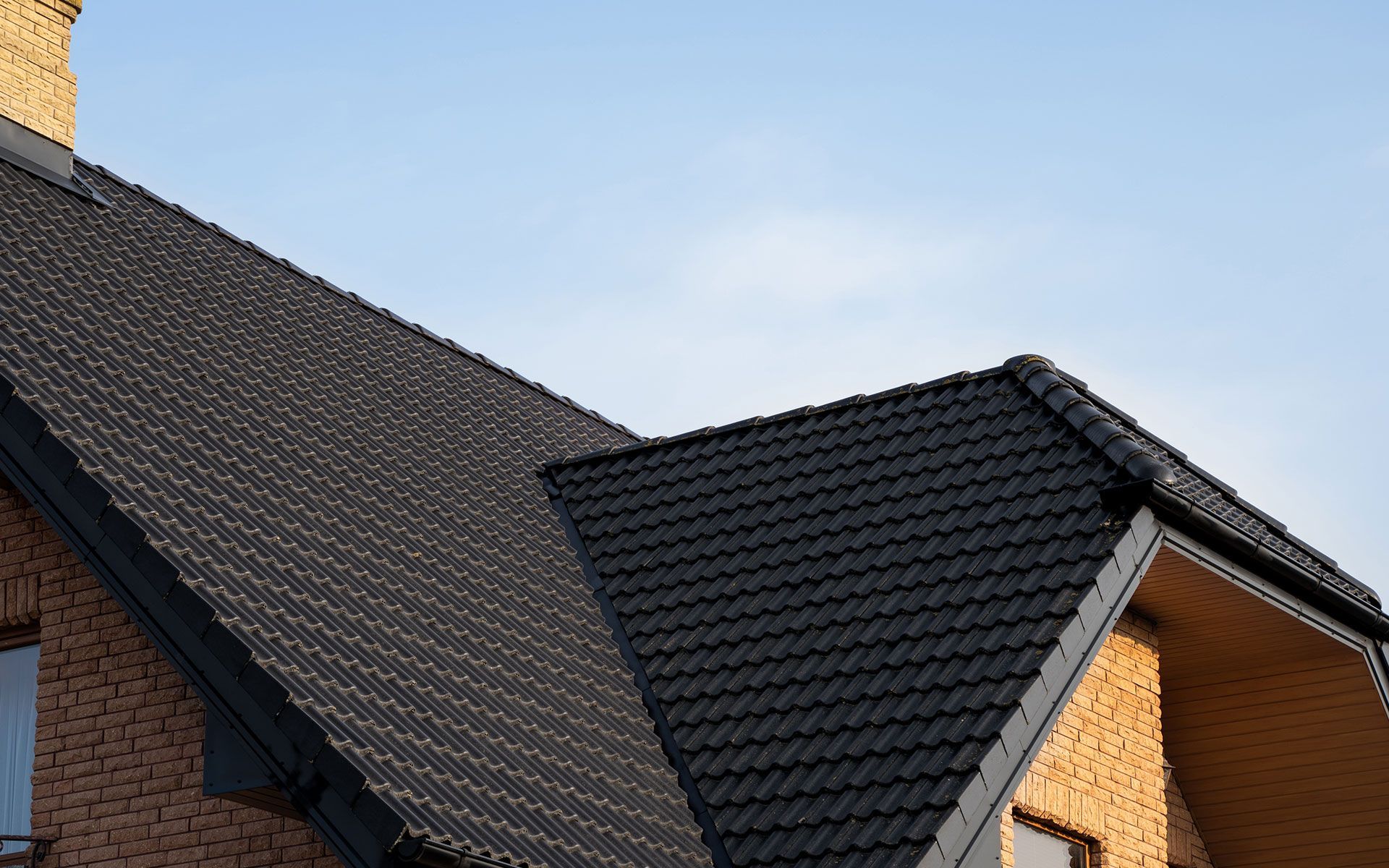 Dark gray tiled roof on a brick house with blue sky background.