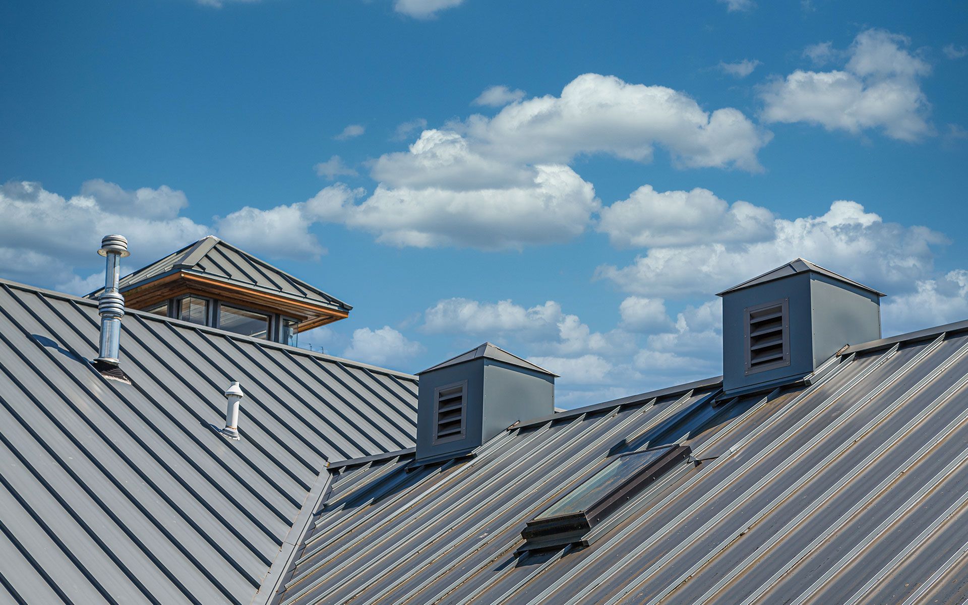 Gray metal roof with vents and a small wooden cupola against a blue sky with fluffy white clouds.