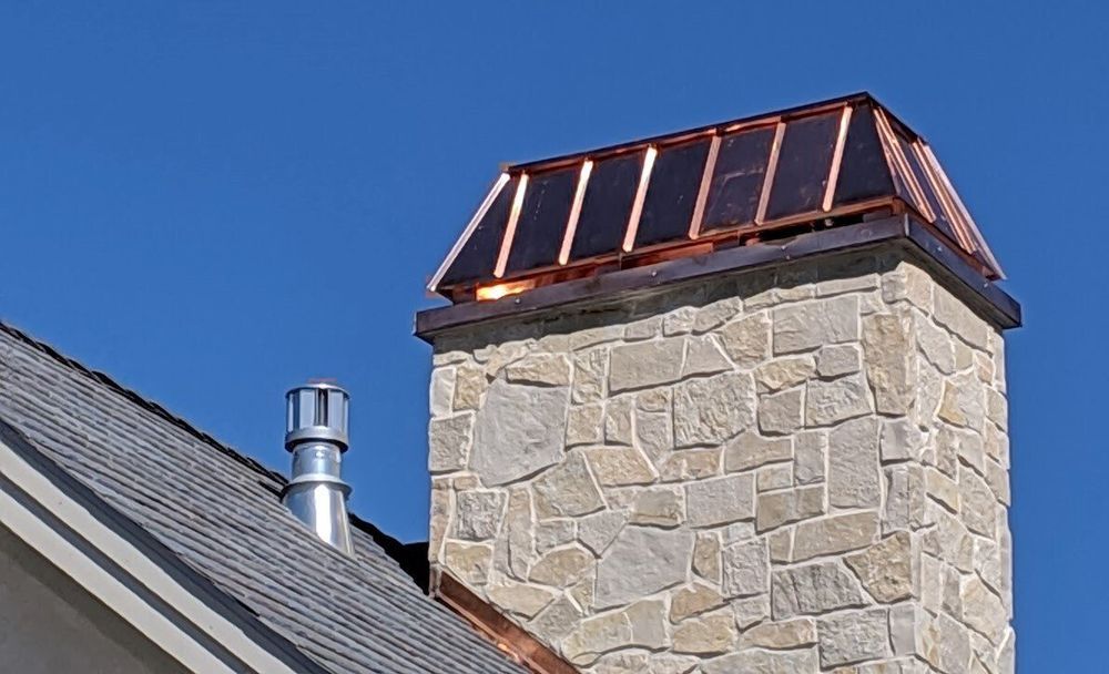 Stone chimney with copper cap against a blue sky, next to a gray roof and metal vent.
