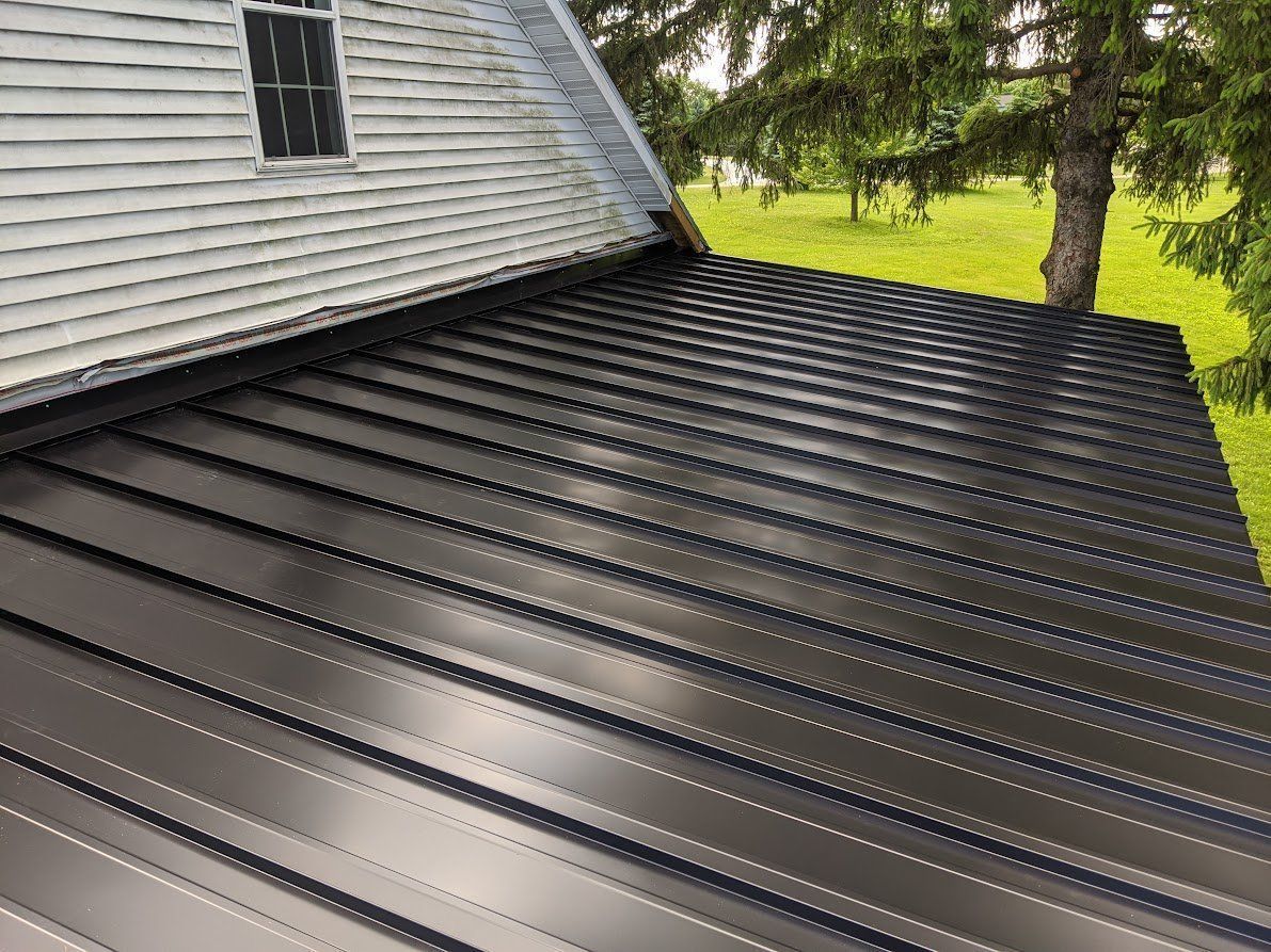 Black metal roof on a house, contrasting with the gray siding and green trees in the background. Black metal roof on a house, contrasting with the gray siding and green trees in the background.