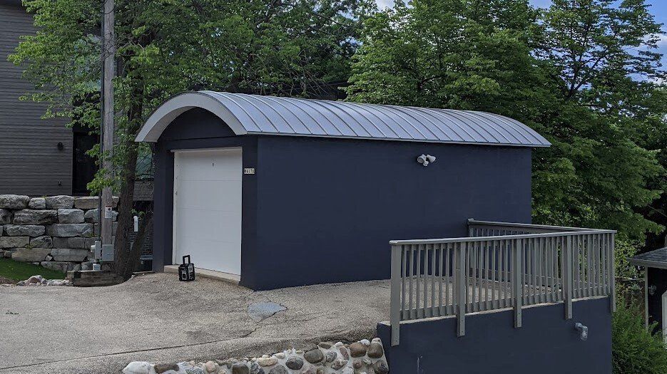 Blue garage with a curved metal roof, white door, and small deck.