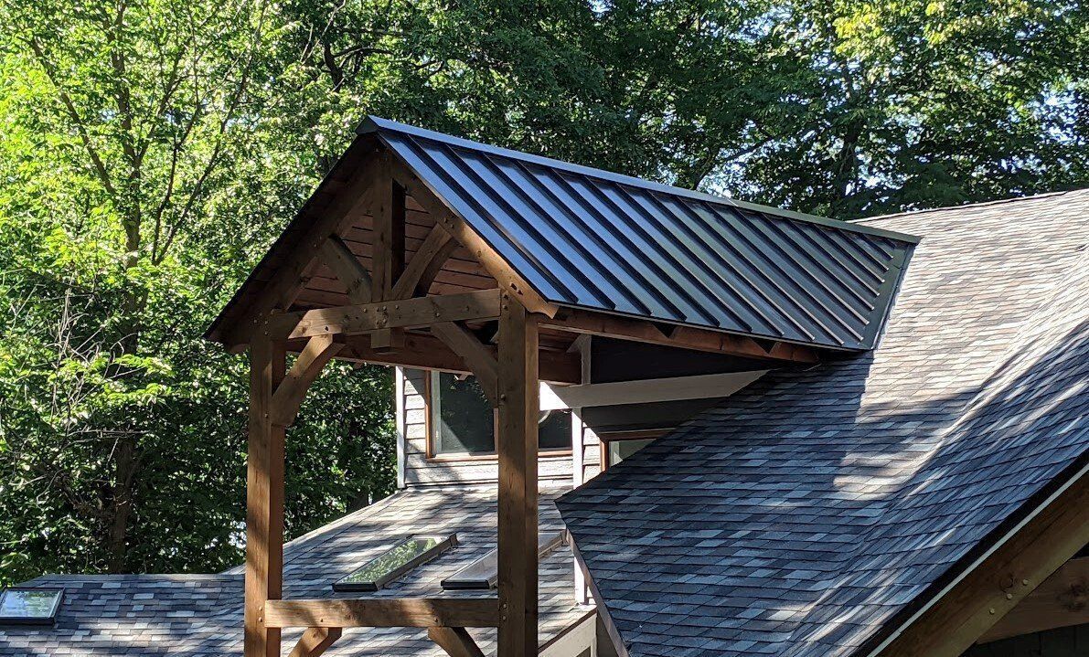 Wooden porch with corrugated metal roof. Shingled roof in foreground, trees in background.